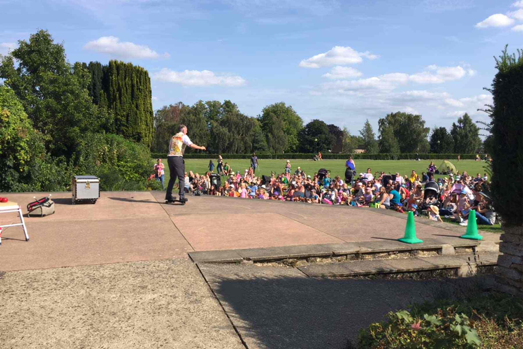 Outdoor Science Show Dr Ken is showing push and pull forces whilst riding snake board in front of an audience sat on the grass in Waterloo park, Norwich.