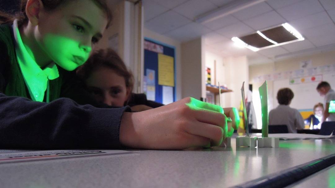 Laser and Light Workshop Pupils deep in concentration whilst attempting to see if light travels in a straight line. They shine a green light through 3 vertically aligned identical pieces of card, each card has small holes strategically placed at the same height.