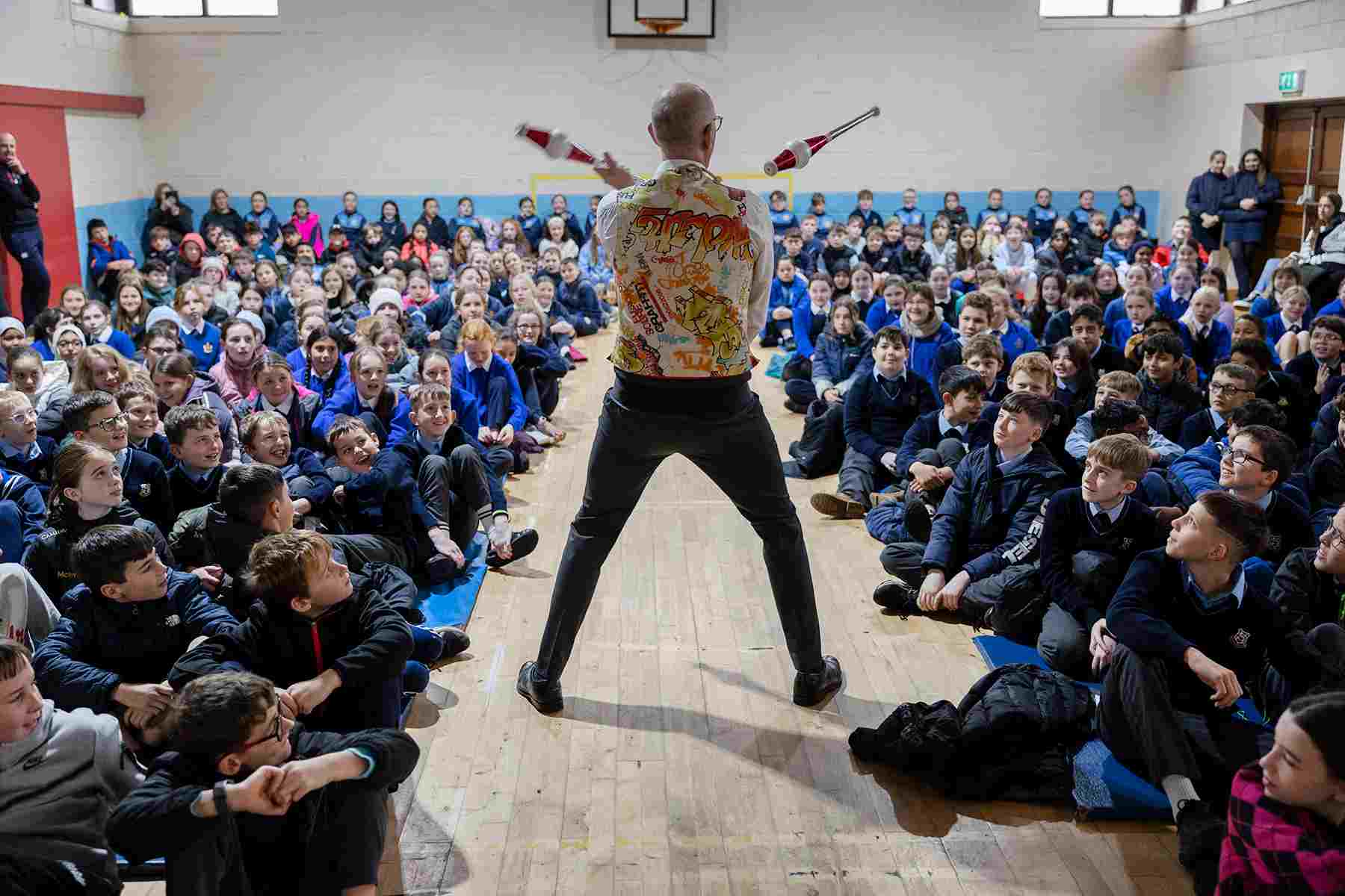 Rear view of Dr Ken juggling clubs in front of seated audience of high school pupils.